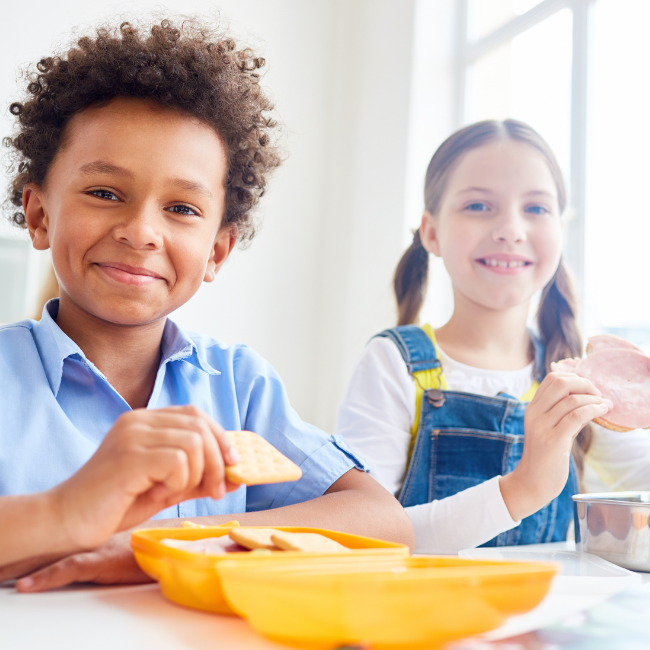 Two children at a table, eating. One has a yellow lunchbox open in front of them.