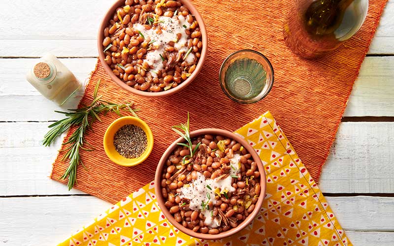 Two peach bowls are filled with Bama Beans & Pork, and garnished with Alabama White BBQ sauce and freshly minced rosemary. 