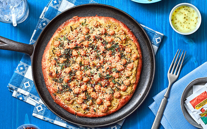 Pesto Garbanzo Pizza served atop a black serving plan and garnished with parmesan cheese, red pepper flakes and oregano. 