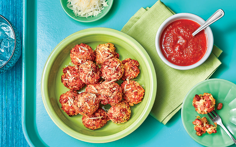 Multiple pieces of Baked Bean Arancini are garnished with Parmesan cheese, and are sitting in a green bowl next to a dish of marinara sauce.