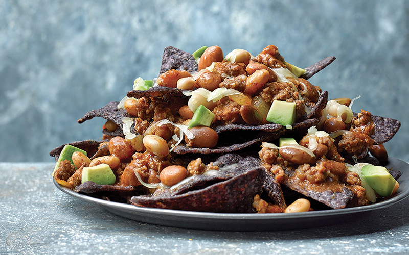 Blue corn chips piled high on a plate with chili, bison, and avocado