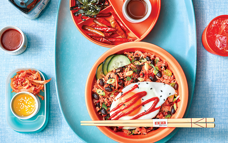 Black Bean Bibimbap in a peach colored bowl with chop sticks topped with an over-easy egg, sliced cucumber, and gochujang ketchup.