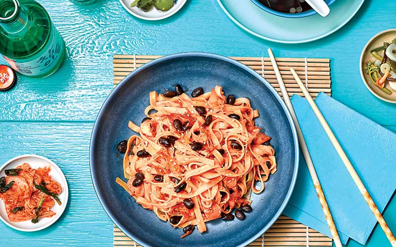Black Bean & Kimchi Noodles is plated with toasted white sesame seeds and scallions, and sits on a blue bowl next to a pair of chopsticks. 