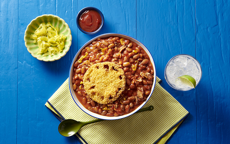 A white bowl with a blue rim is filled with Carolina Cottage Chili topped with a cornmeal-dusted fried tomato slice.