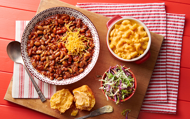Memphis BBQ-Style Chili is filling a white bowl with red decals and served with a small bowl of mac and cheese, coleslaw, and cornbread. 