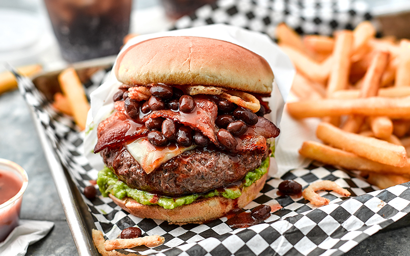 Ole fiesta bean burger sits on checkered serving paper in front of french fries.
