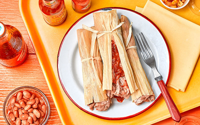Three tamales wrapped in corn husks on a plate on a yellow tray.