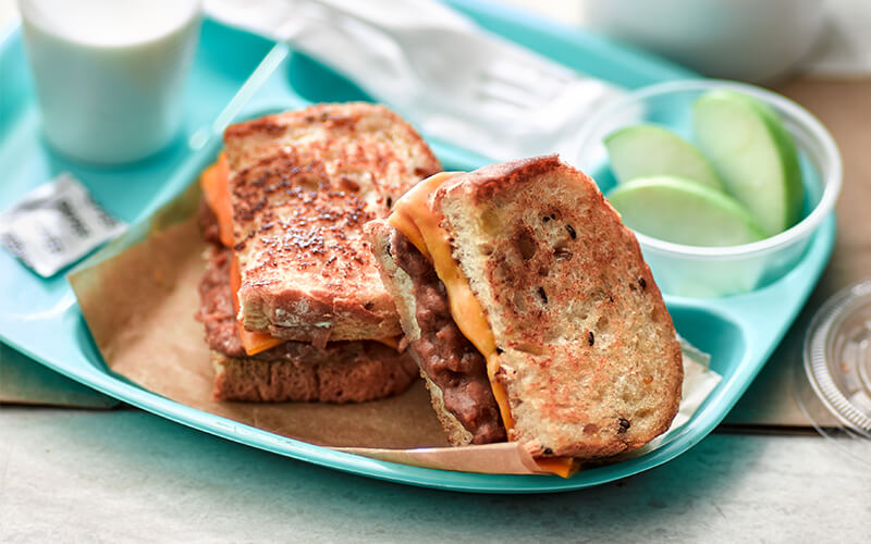 Blue tray holding a glass of milk, a grilled cheese with beans sliced in half, and apple slices