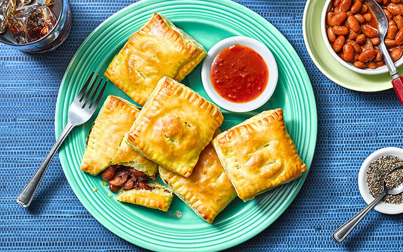 Golden brown Reggae Falls Hand Pies stacked atop a bright blue plate and served with a dish of red dipping sauce. 