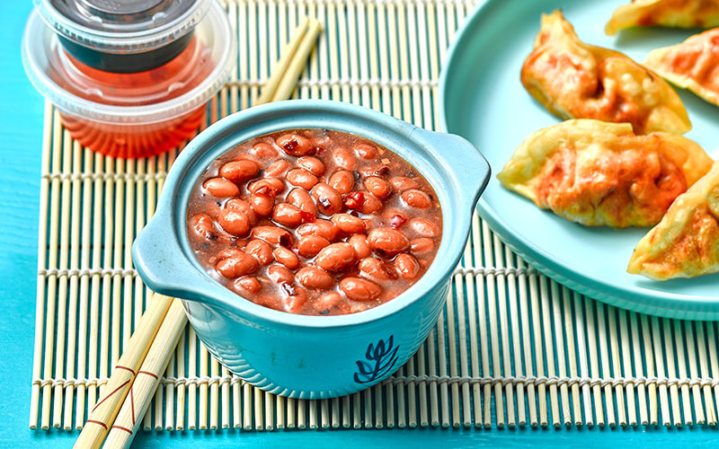 Small bowl of sweet chili baked beans on a bamboo mat with chopsticks, soy sauce, and a plate of potstickers