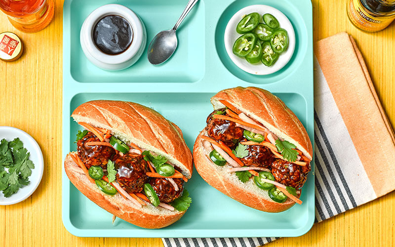 Two Veggie Bahn Mi Meatball sandwiches topped with jalapeño, hoisin sauce, and pickled vegetables served atop a blue tray.