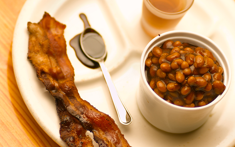 ramekin of apple bourbon baked beans on a divided plate next to two strips of crispy bacon