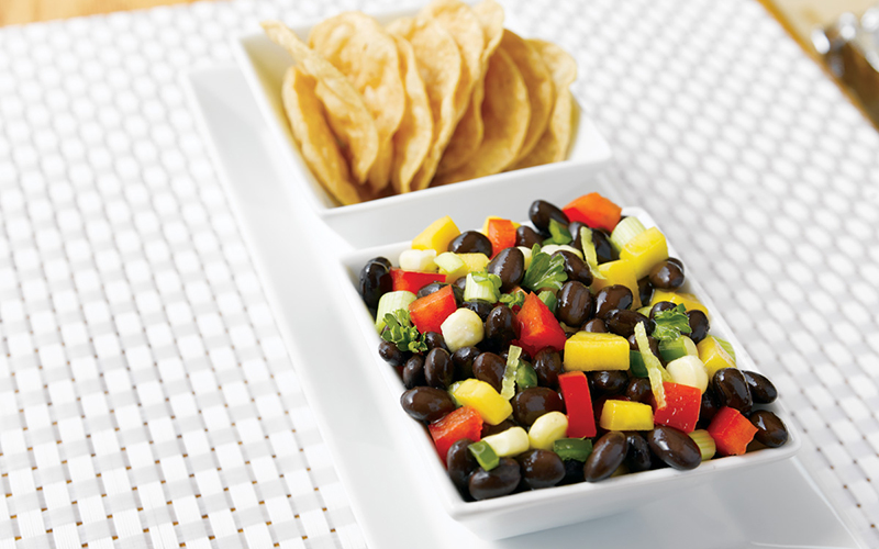 Two small white square bowls, the front one holds black bean mango salsa and the back one holds tortilla chips