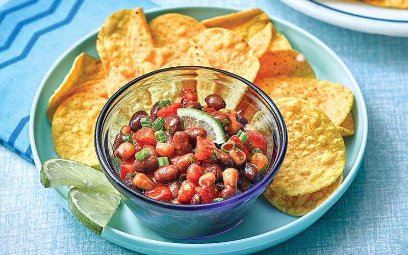 A clear blue bowl is filled with Bright Bean Salsa and served next to round tortilla chips along with two lime slices. 