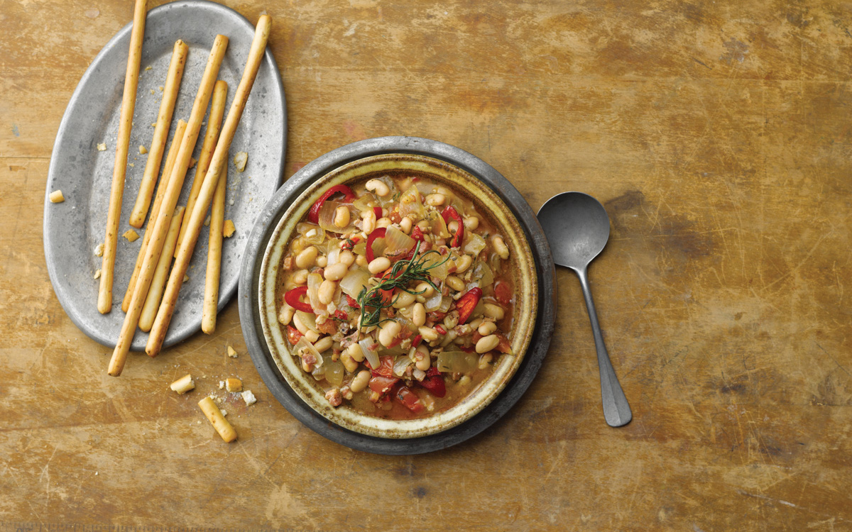 Earthenware plate with white beans, pancetta, and onions topped with fried rosemary and red chiles