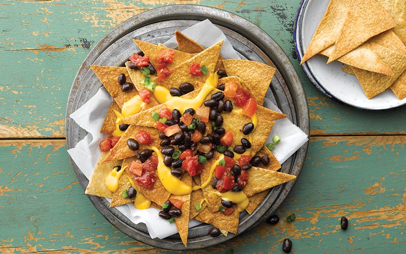 Plate of crispy baked tortillas topped with beans, tomatoes, and garbanzo nacho cheese sauce