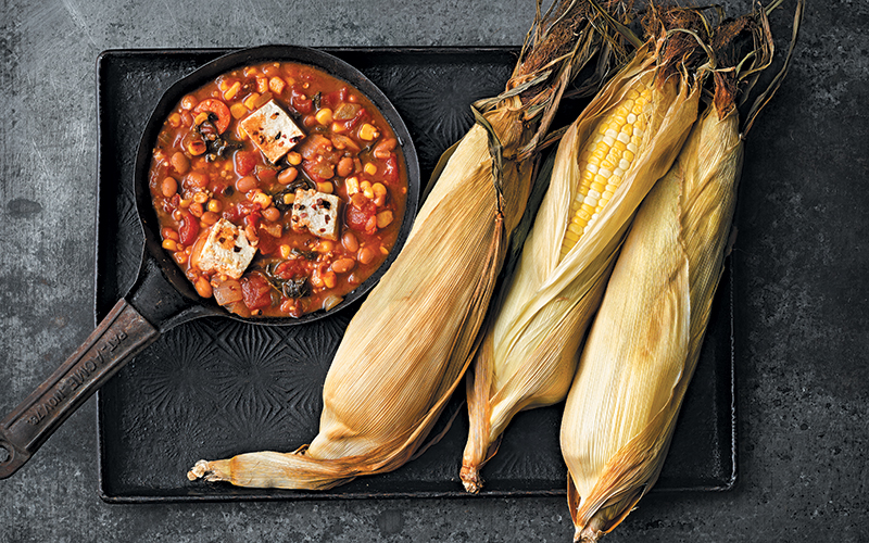 A small cast iron pan holds vegetarian baked bean chili with tofu and kale, next to three ears of roasted corn
